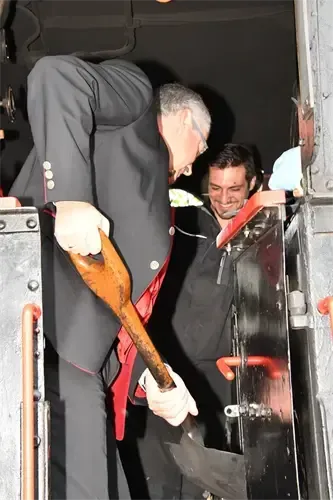 Ein Mann in formeller Kleidung hält eine Axt, während ein anderer Mann neben ihm lächelt, sie sind in der Nähe einer Tür.