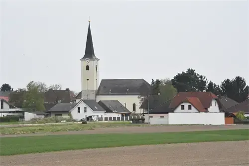 Eine weiße Kirche mit einem Kirchturm steht inmitten mehrerer Wohngebäude. Die Kirche ist von Bäumen umgeben, und im Vordergrund befindet sich ein Grasfeld.