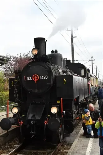 Eine schwarze Vintage-Lokomotive mit der Bezeichnung 93 und 1420, aus der Rauch austritt, steht auf den Gleisen, während sich Menschen darum versammeln.