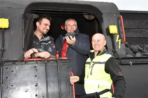 Drei Männer posieren mit einem Kohlestück in einem Kohlezug, einer trägt eine Brille, ein anderer eine gelbe Weste.