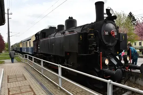 Eine schwarze Dampflokomotive steht auf einem Bahngleis mit einer Metallbrüstung. Es gibt Menschen auf dem Bahnsteig, und einige sind in der Nähe des Zuges. Es gibt ein Gebäude mit einem Zaun und einem Baum in der Ferne.