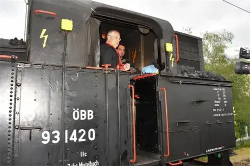 Zwei Männer in einem Zug mit 'OeBB 931420' auf der Seite. Ein Mann mit Brille schaut aus dem Fenster.