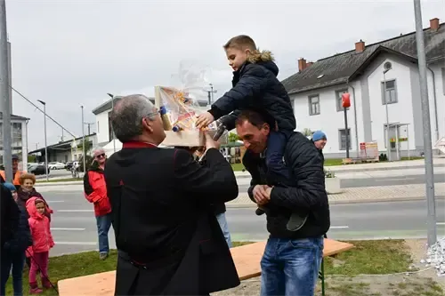 Ein erwachsener Mann hält ein Geschenk, während ein Junge auf seinen Schultern sitzt, umgeben von Menschen auf einer Rasenfläche in der Nähe der Straße.