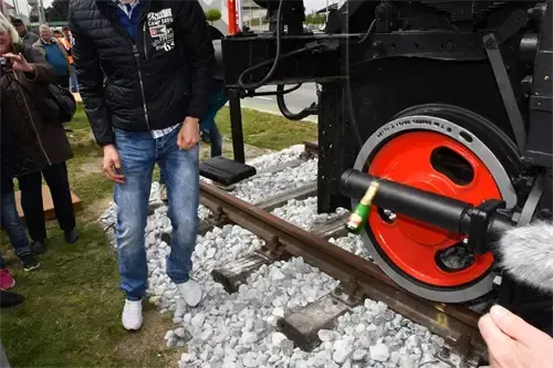 Ein Mann geht auf einem Bahngleis mit einem Mikrofon und einer Flasche in der Nähe eines Zugrades. Hinter ihm beobachten andere. Die Umgebung ist im Freien mit Kies und Gras.