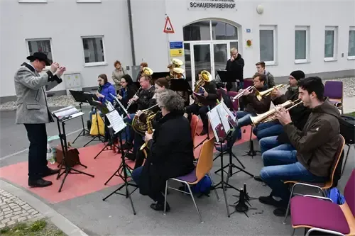 Eine Brass-Band übt auf der Straße vor einem weißen Gebäude, mit sitzenden und stehenden Musikern und einem Dirigenten vorne.
