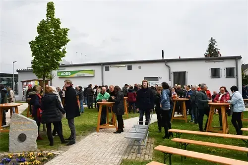 Eine Gruppe von Menschen steht um Tische in einem Grasbereich vor einem Gebäude mit einem Schild, das 'ADEG' lautet. Bäume und Blumen schmücken die Umgebung.