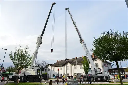 Zwei Kräne sind vor einem weißen Gebäude positioniert und heben ein Objekt. Eine Menschenmenge steht unten und beobachtet. Im Hintergrund sind Bäume und Straßenlaternen zu sehen.