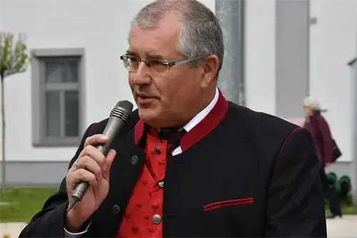 Ein älterer Mann in einem roten Vest und einem schwarzen Anzug spricht in ein Mikrofon vor einem weißen Gebäude mit Fenstern und einem Baum im Vordergrund.