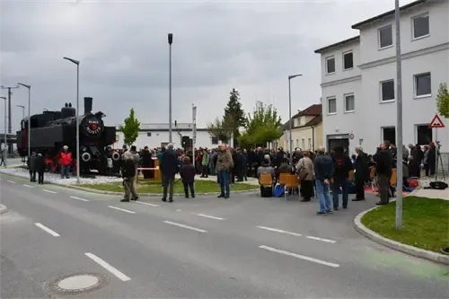 Eine große Menschenmenge versammelt sich um eine alte Dampflokomotive, die vor einem Gebäude geparkt ist. Die Leute stehen und sitzen auf Stühlen, manche tragen Hüte.