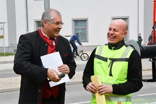Zwei Männer stehen in einer urbanen Umgebung, einer in formeller Kleidung hält ein Papier und ein Mikrofon und lächelt. Der andere trägt eine Warnweste und hält eine gelbe Mappe, mit einem Radfahrer im Hintergrund.
