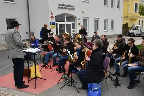 Eine Gruppe von Musikern spielt Instrumente auf einer Bühne vor einem Gebäude mit dem Schild WOHNHAUSANSLAGE SCHAURHUBERHOF. Ein Mann in einem Hut dirigiert.