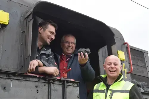 Drei Männer stehen in einem Zugabteil. Der Mann in der Mitte hält einen Felsen, während der Mann auf der linken Seite sich an das Fenster lehnt und der Mann auf der rechten Seite lächelt.
