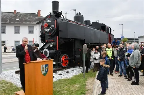 Ein Mann spricht an einem Podium vor einer schwarzen Vintage-Lok mit 'ORTTZ' auf der Front. Eine Menschenmenge steht auf einem Bürgersteig. Ein Gebäude mit vielen Fenstern befindet sich hinter der Lok.