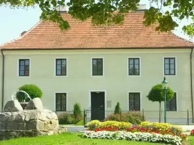 Ein großes weißes Gebäude mit rotem Dach, umgeben von üppigem Grün, hat mehrere Fenster und einen Garten mit bunten Blumen und einem Brunnen davor.