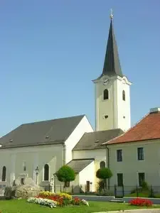 Eine weiße Kirche mit einem Kirchturm und Uhren, umgeben von einem kleinen Garten mit Blumen und einem kleinen Weg, der zum Eingang führt.