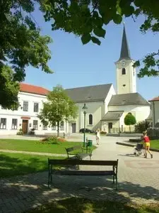 Ein Außenbereich mit einer Kirche, spazieren gehende Menschen und einer Bank vor einer Rasenfläche mit Bäumen und klarem Himmel.