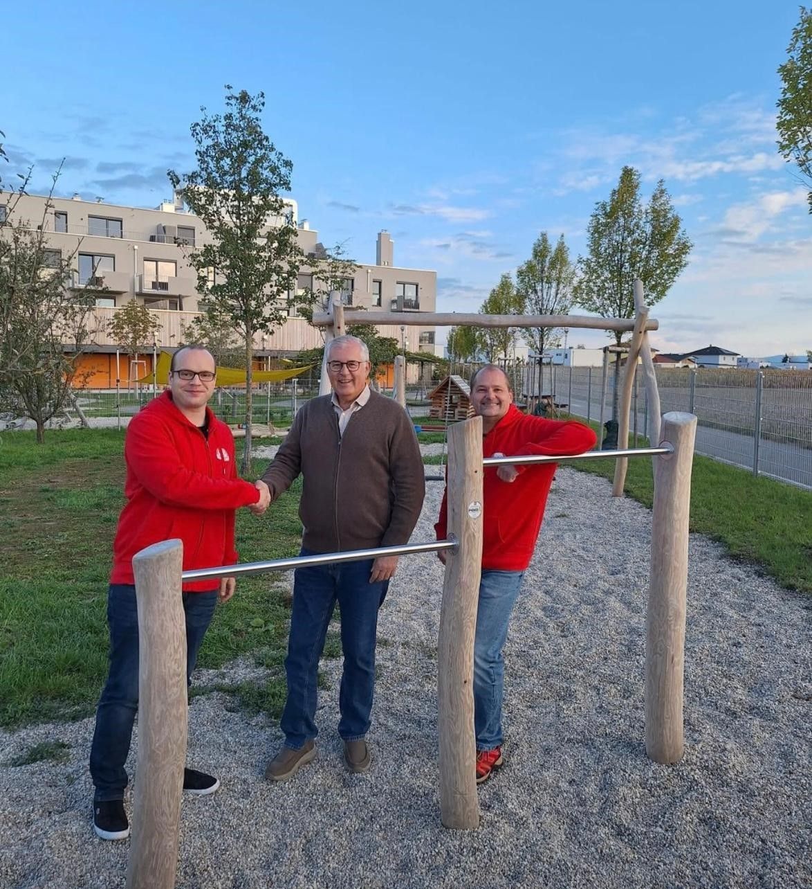 Drei Männer in roten Jacken stehen lächelnd an einem Outdoor-Fitnessstudio. Dahinter ist ein Spielplatz zu sehen. Ein Gebäude ist im Hintergrund und ein Zaun verläuft entlang der Seite.