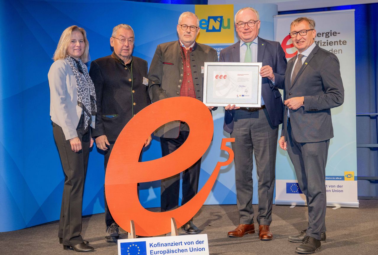 Fünf Erwachsene stehen hinter einem großen orangefarbenen E5-Logo und halten eine Urkunde in der Hand. Sie befinden sich in einem Konferenzsaal mit blauer Wand und einer EU-Banner.