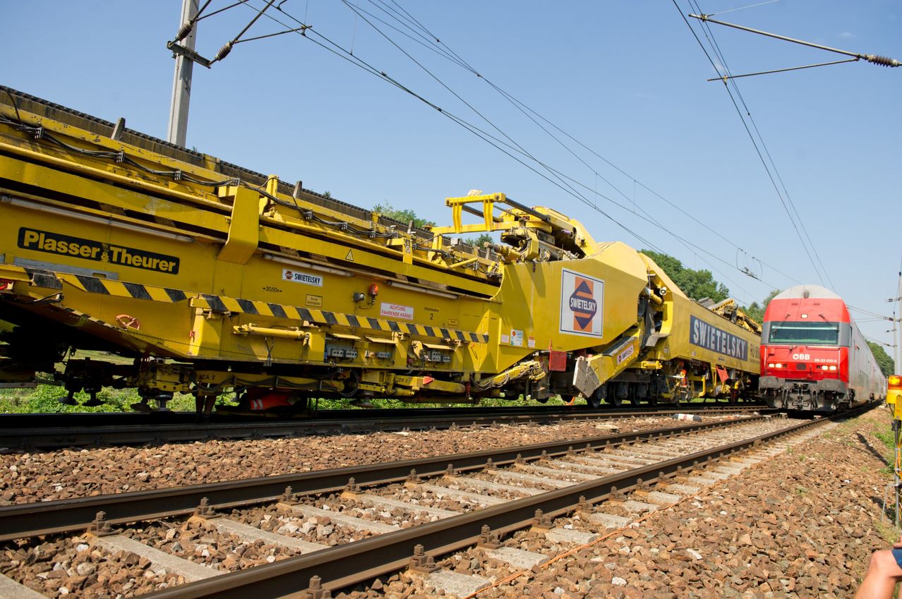 Ein gelber Bauzug steht auf den Bahngleisen mit Kies an den Seiten. Er hat mehrere Abschnitte, auf denen der Name Swietelsky deutlich zu sehen ist.