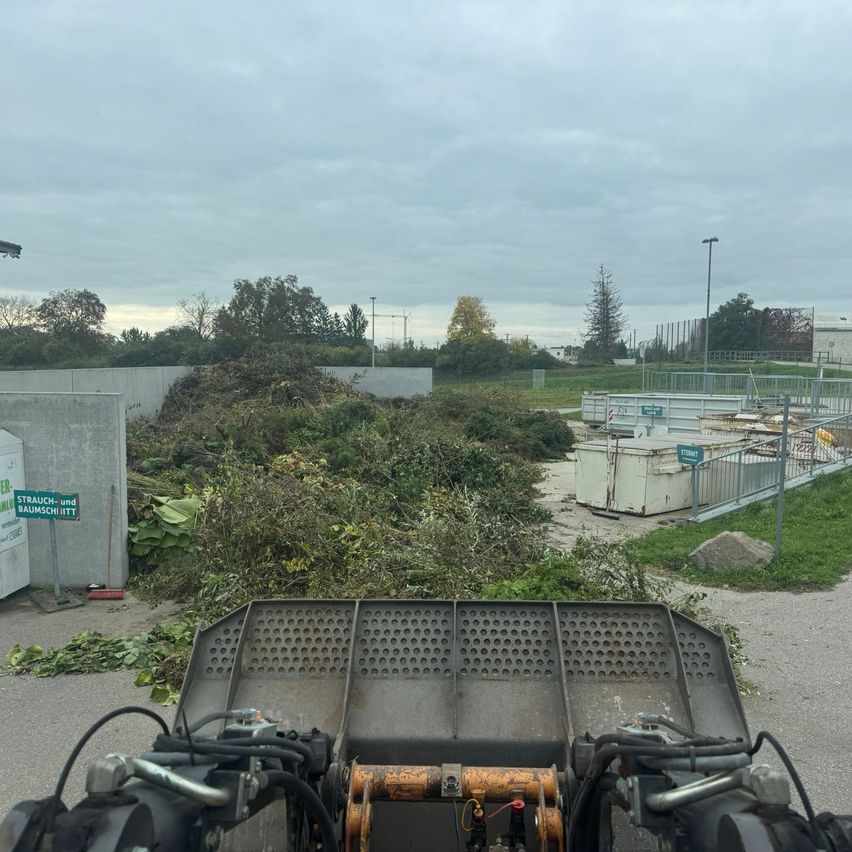 Ein Blick von einem Maschinenfahrzeug zeigt eine Landschaft mit einem großen Haufen geschnittener Sträucher und Bäume, einem eingezäunten Bereich und einem bewölkten Himmel.