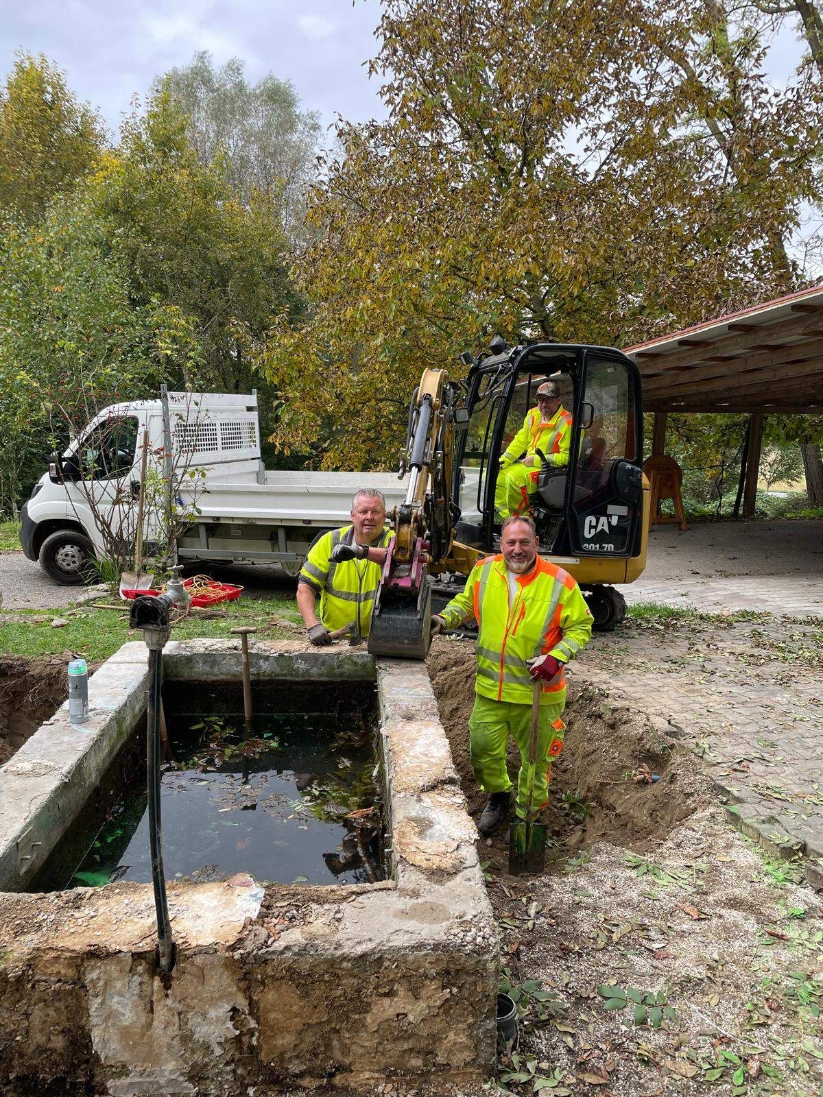 Drei Arbeiter in gelber Sicherheitskleidung stehen draußen neben einem großen Loch. Ein CAT-Bagger steht hinter ihnen.