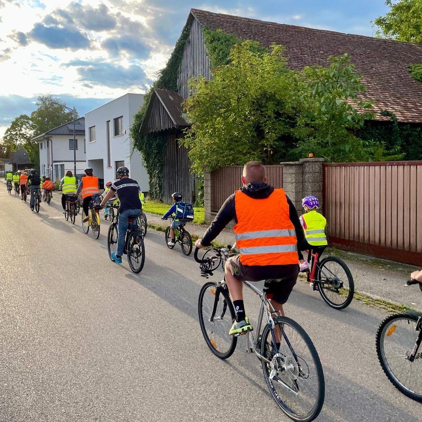 Eine Gruppe Radfahrer fährt eine Straße entlang, mit Erwachsenen in Sicherheitswesten und Kindern mit Helmen. Gebäude und Bäume säumen die Straße, und der Himmel ist teilweise bewölkt.
