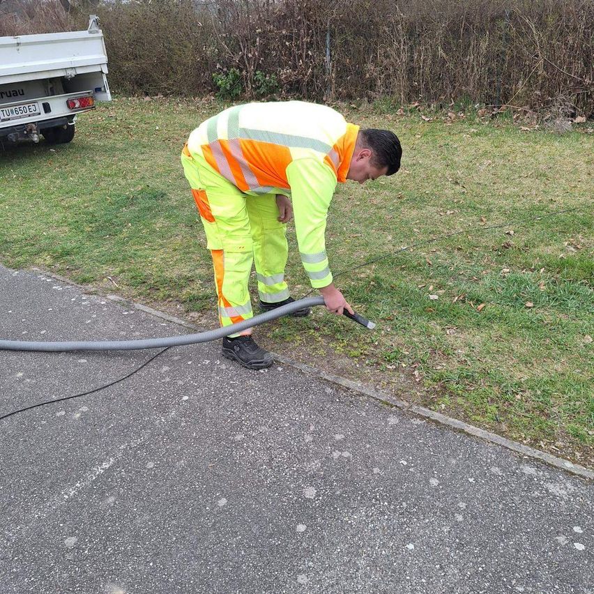 Ein Mann in einer Warnweste wässert mit einem Schlauch das Gras am Straßenrand. Ein weißer Lkw ist in der Nähe geparkt.