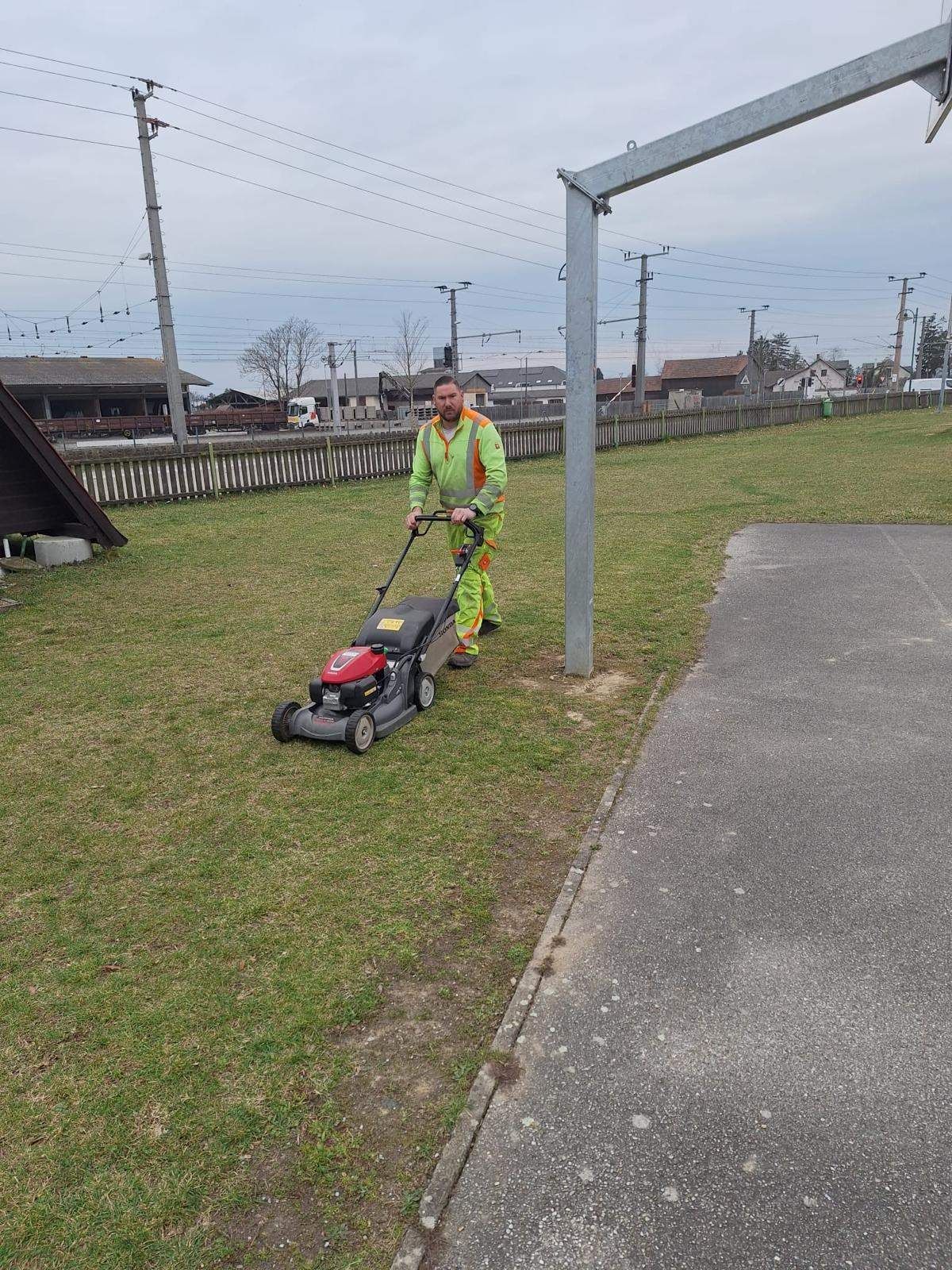 Ein Mann in Sicherheitsausrüstung mäht den Rasen in der Nähe eines Metallpfahls. Das Gras ist üppig und grün. Im Hintergrund befinden sich ein Zaun, Häuser und Strommasten.
