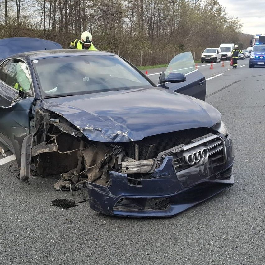 Ein blauer Audi wurde auf der Straße zerstört. Rettungsfahrzeuge sind anwesend, und Arbeiter kümmern sich um den Unfallort.