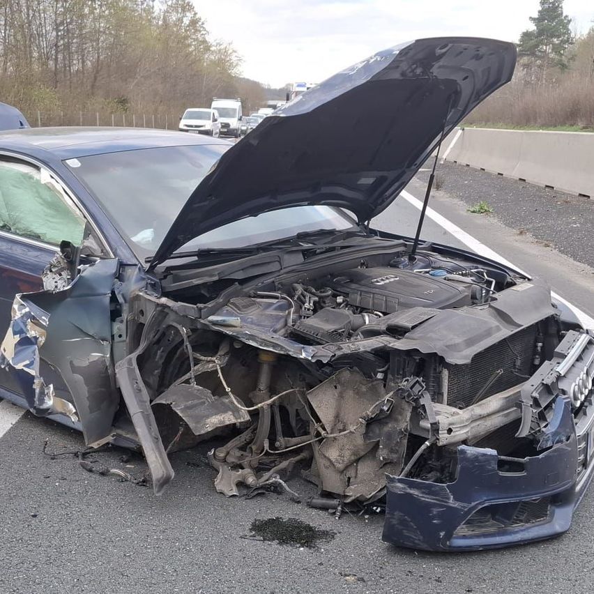 Ein stark beschädigtes blaues Auto steht am Straßenrand mit offenem Motorhaube und zeigt erhebliche Schäden an der Front.