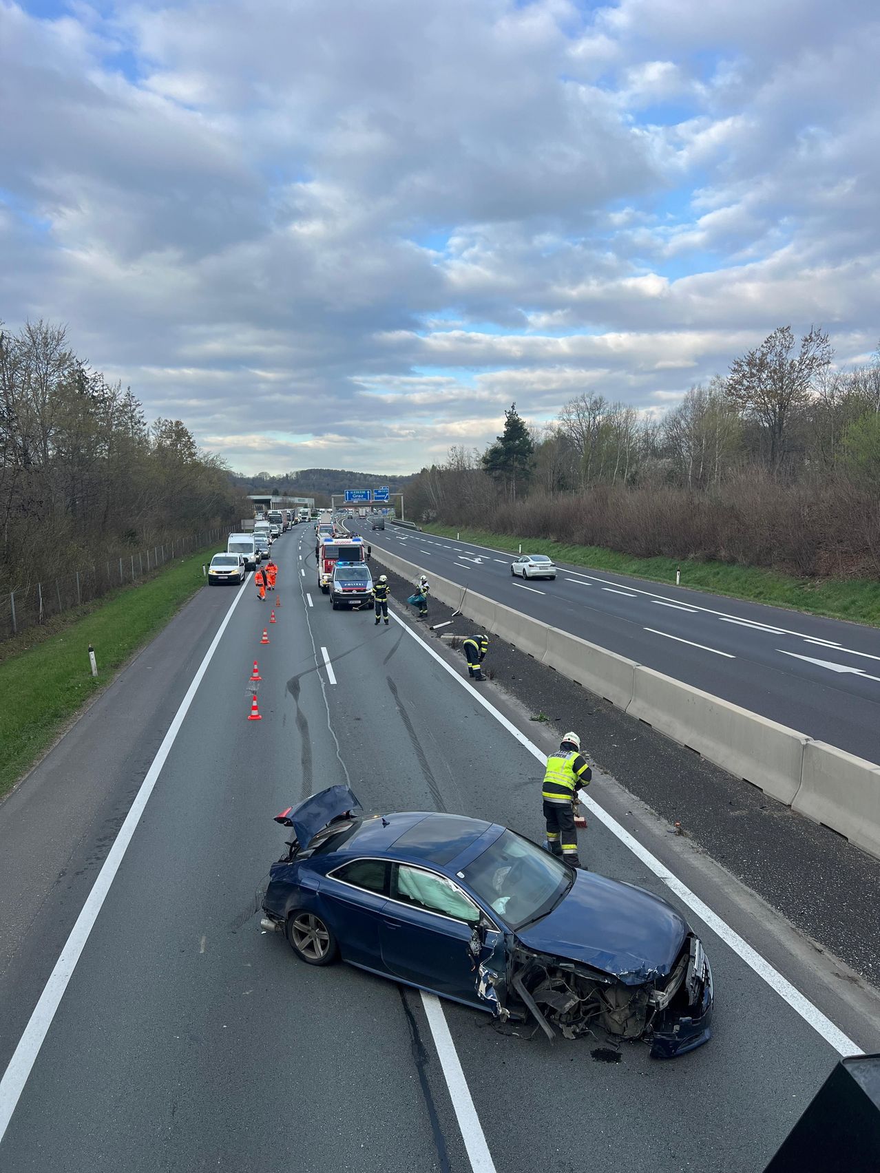 Ein blaues Auto ist auf einer Autobahn verunglückt. Arbeiter in gelben Westen und Helmen markieren den Bereich mit orangefarbenen Kegel.