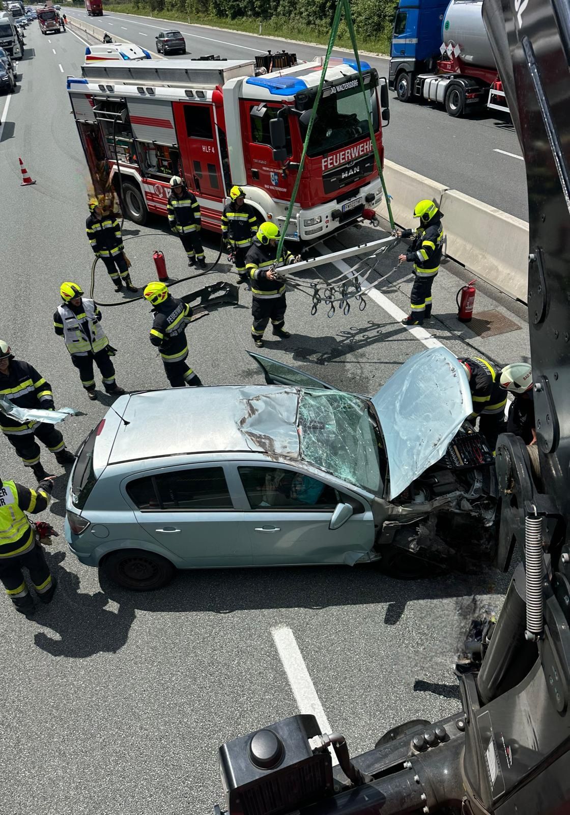 Feuerwehrleute kümmern sich um ein beschädigtes Fahrzeug auf der Straße. Die Front des Autos ist zerquetscht und die Motorhaube ist offen. Mehrere Feuerwehrleute sind um das Auto herum.