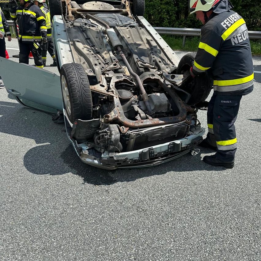 Feuerwehrleute untersuchen ein umgekipptes Auto am Straßenrand. Der Unterboden des Autos ist freigelegt und ein Rad ist abmontiert. Ein Feuerwehrmann untersucht das Fahrzeug.