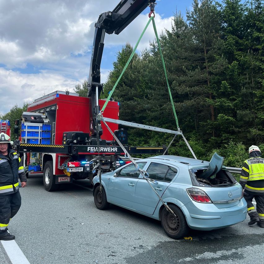 Ein Kran hebt ein beschädigtes blaues Auto, mit Feuerwehrleuten und einem Verkehrsleiter in der Nähe. Das Dach des Autos ist ab und es liegt auf der Straße.