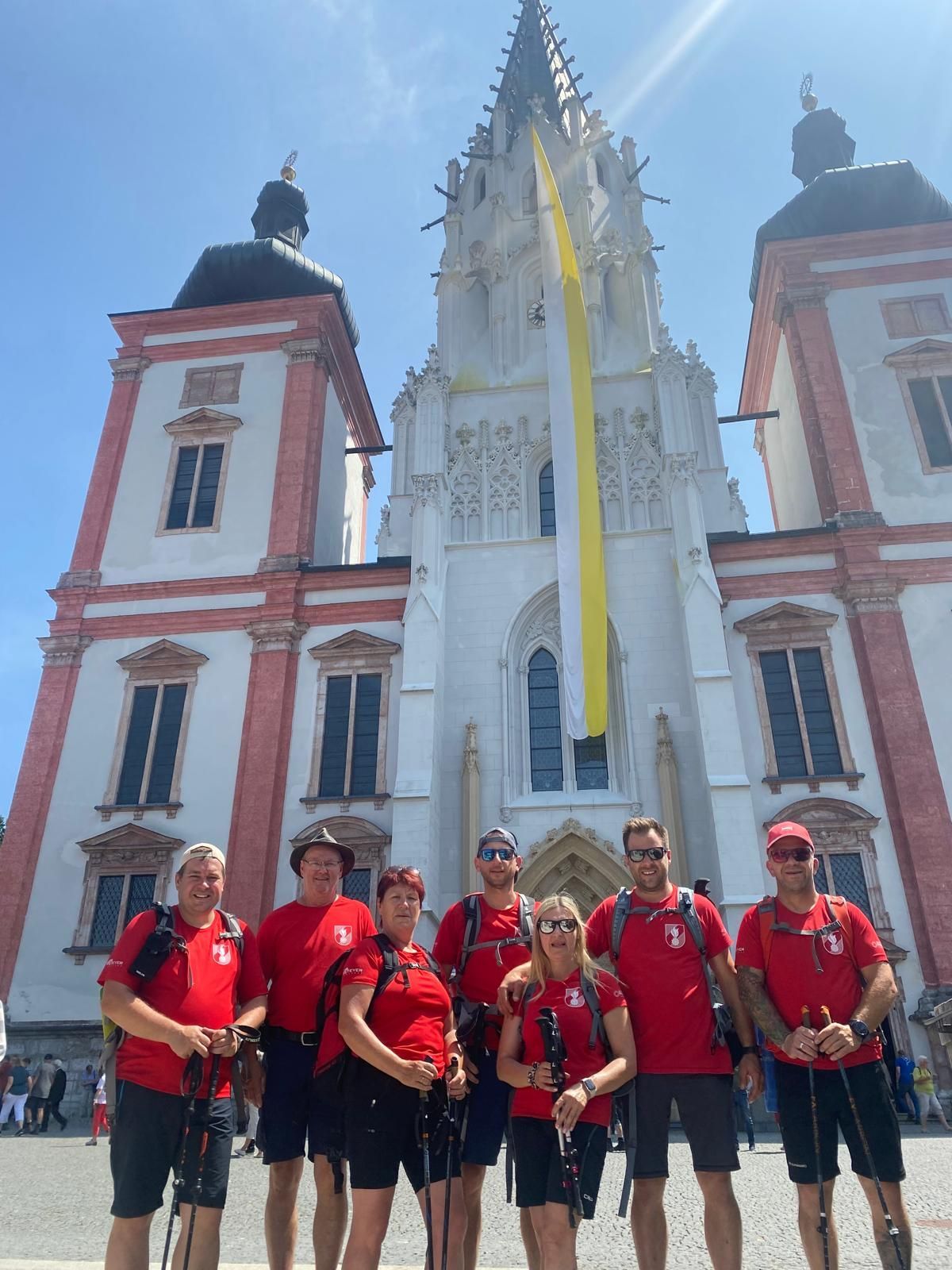 Eine Gruppe von Menschen in roten Shirts und Hüten posiert vor einer großen weißen und roten Kathedrale unter einem klaren blauen Himmel.