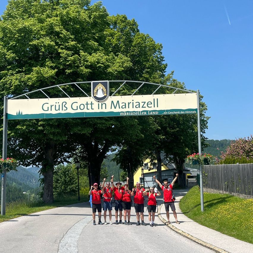 Eine Gruppe von Menschen in roten Shirts und schwarzen Shorts steht unter einem Schild mit der Aufschrift Gruß Gott in Mariazell. Sie lächeln und heben ihre Hände. Dahinter stehen Bäume, ein Zaun und ein gelbes Haus. Der Himmel ist klar und blau.