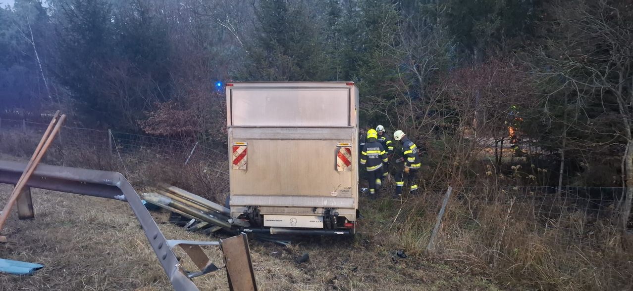 Drei Feuerwehrleute in gelben Uniformen inspizieren einen beschädigten LKW am Straßenrand, umgeben von Bäumen und Trümmern.