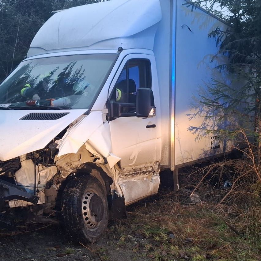 Ein weißer Lieferwagen ist auf einer Straße demoliert, mit stark beschädigtem Frontstoßfänger und Motorhaube. Rettungskräfte sind anwesend.