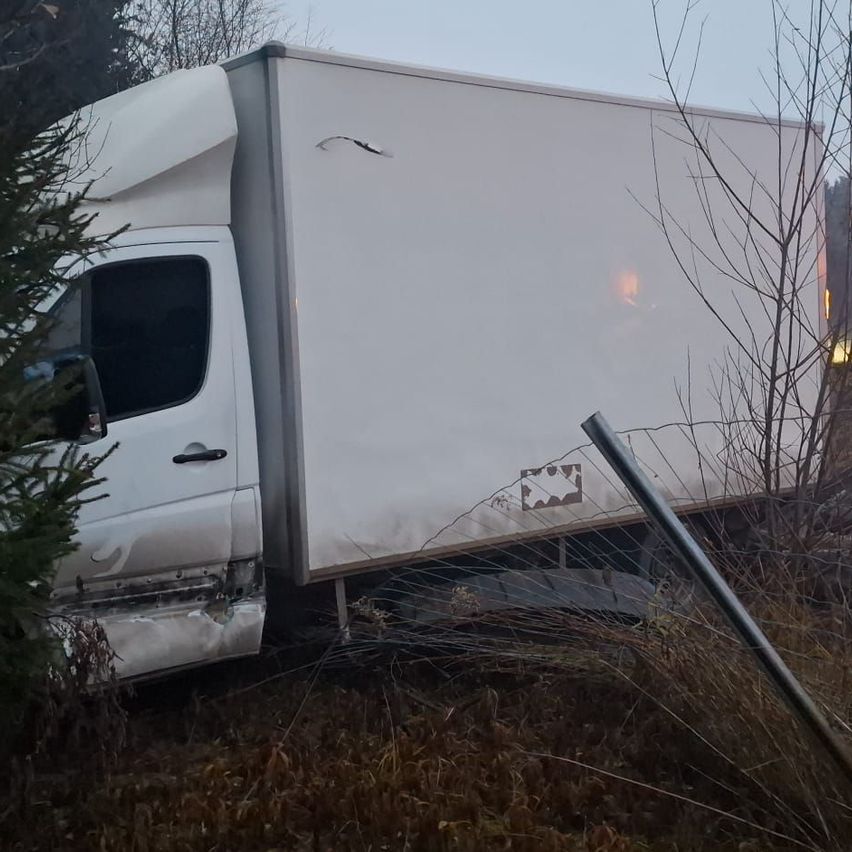 Ein weißer Lieferwagen mit beschädigter Heckpartie steht abseits der Straße, umgeben von trockenem Gras und einem zerbrochenen Pfosten.