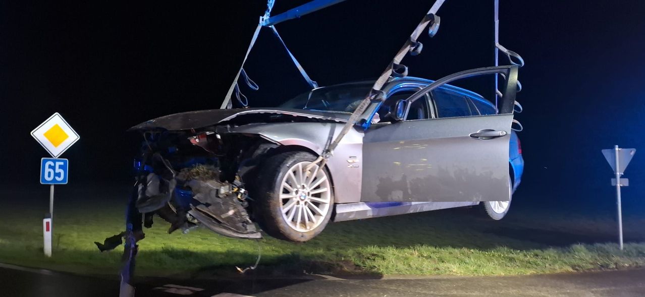 Ein schwer beschädigter BMW-Wagen wird bei Nacht von einem Kran angehoben. Die Front ist zerknittert und das Rad hängt herunter. Das Auto steht auf Gras.