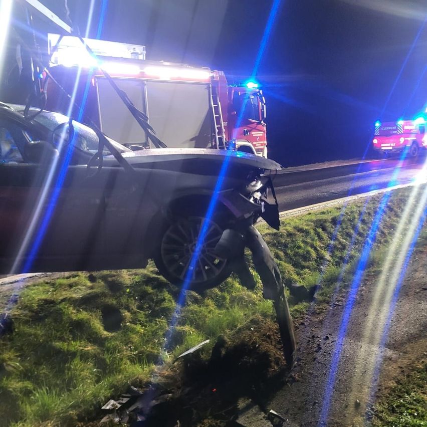 Ein Autounfall mit Einsatzfahrzeugen am Straßenrand bei Nacht. Ein Auto steht abseits der Straße, seine Front ist schwer beschädigt.