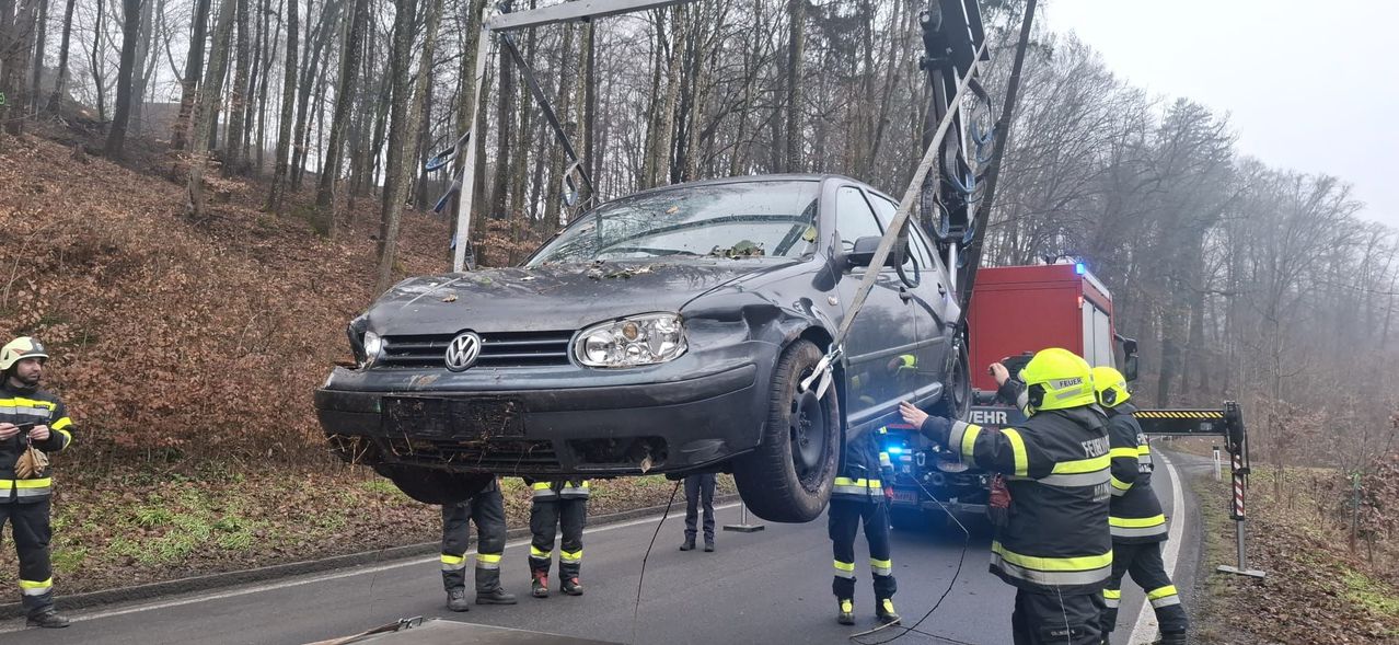 Feuerwehrleute heben einen beschädigten Volkswagen Golf mit einem Kran von der Straße. Das Auto ist an Seilen aufgehängt und der Stoßfänger hängt herunter. Es weist sichtbare Frontschäden auf.