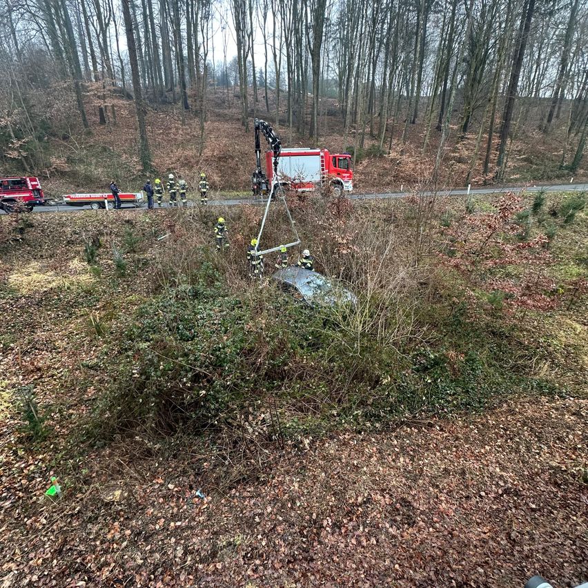 Feuerwehrleute in Schutzkleidung arbeiten daran, ein beschädigtes Fahrzeug aus einem bewaldeten Gebiet zu bergen. Ein roter Feuerwehrwagen ist in der Nähe geparkt.