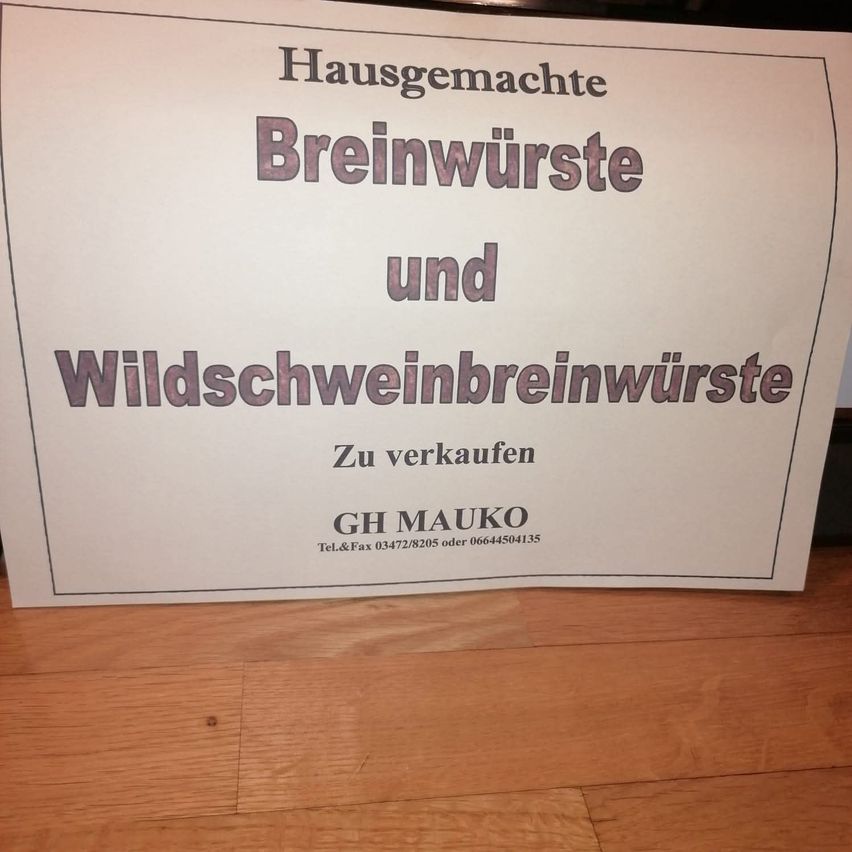 Ein weisses Schild auf einem Holzboden zeigt 'Hausgemachte Breinwürste und Wildschweinbreinwürste Zu verkaufen' in Lila und Schwarz. Unten steht 'GH MAUKO' mit Kontaktinformationen.