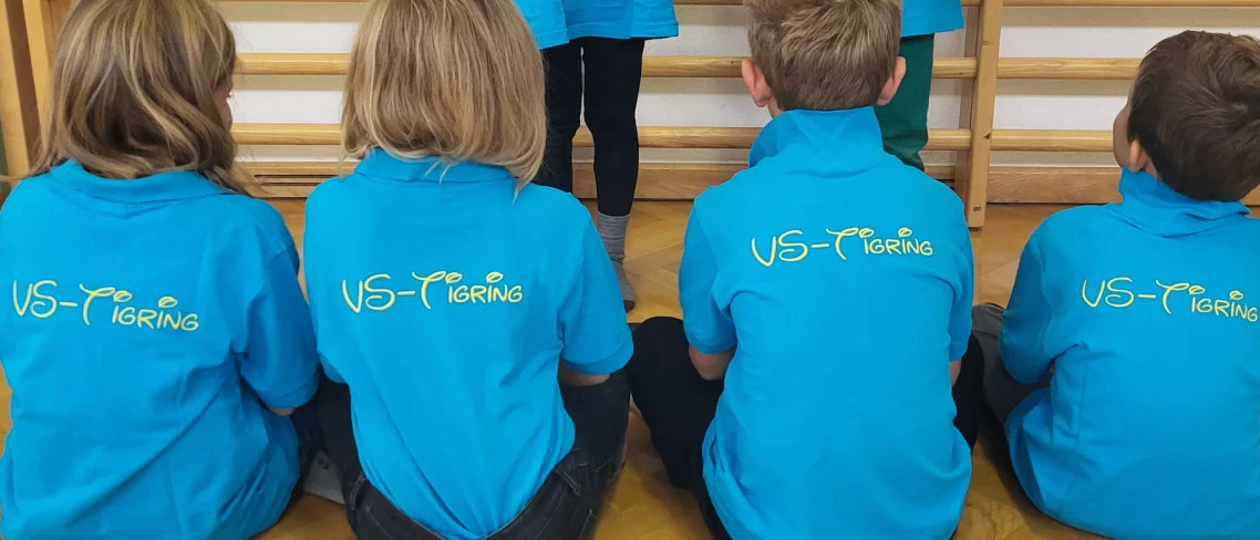 Fünf Menschen sitzen auf dem Boden, drei vorne und zwei stehen dahinter. Sie tragen blaue Shirts mit dem Text 'VS-Tigring'. Der Raum hat Holzböden und im Hintergrund hölzerne Stangen.