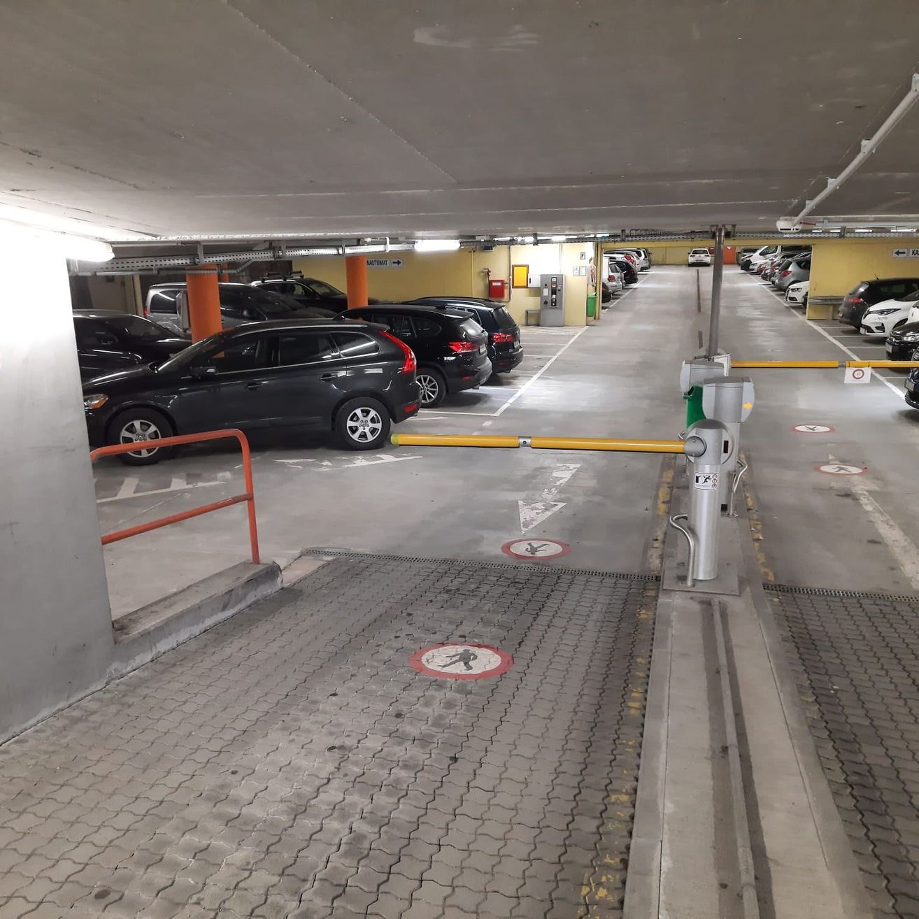 Eine Tiefgarage mit mehreren geparkten Autos. Ein gelber Sperrbalken blockiert den Eingang. Bodenmarkierungen und Schilder sind sichtbar, zusammen mit Lichtern und Säulen.