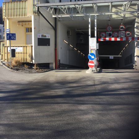 Ein Parkhauseingang mit überdachtem Bereich, Verkehrszeichen und einem Gebäude mit Balkon und Mülleimer in der Nähe.