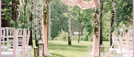 Eine Hochzeitszeremonie im Wald mit weißen Stühlen, Blumendekorationen und einem hölzernen Bogen. Rosa Vorhänge und Laternen schmücken die Szene.