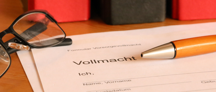 Ein Stift, eine Brille und ein Dokument mit der Aufschrift 'Vollmacht' liegen auf einem Schreibtisch mit rotem Hintergrund. Es gibt einen Slogan, der 'Vorsorge Vollmacht. Rechtzeitig Selbst Bestimmen' lautet. Unten befindet sich ein Logo und die Worte 'Wir sind, wo Sie sind.'