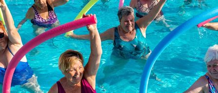 Eine Gruppe von Frauen schwimmt in einem Pool mit bunten Übungsgeräten. Sie lächeln und scheinen es zu genießen.